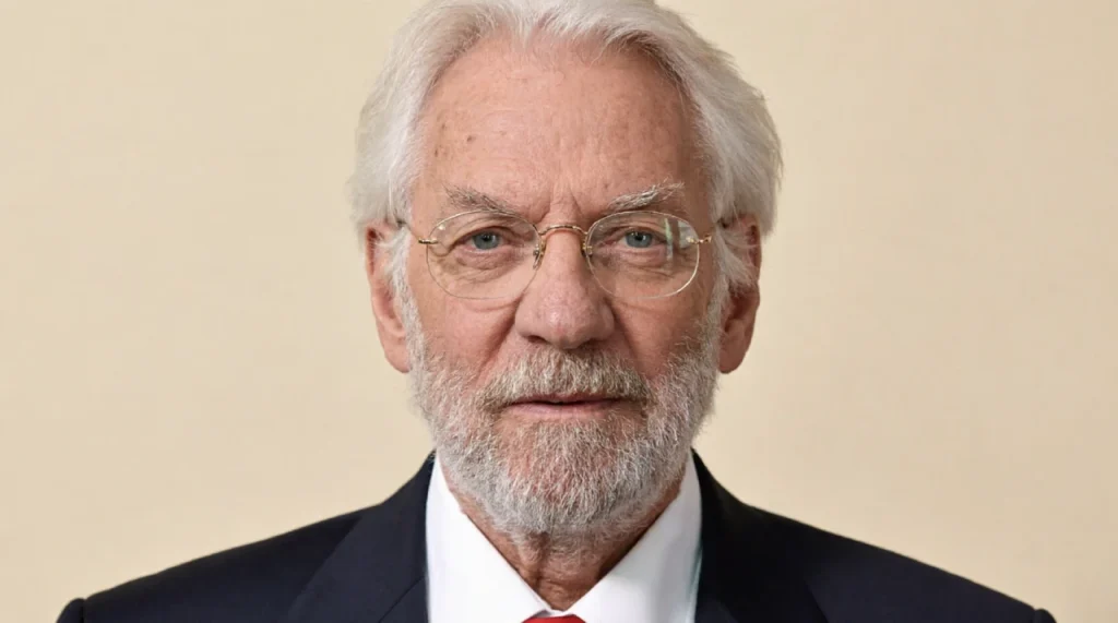 Donald Sutherland with white beard, intense gaze, dark coat.