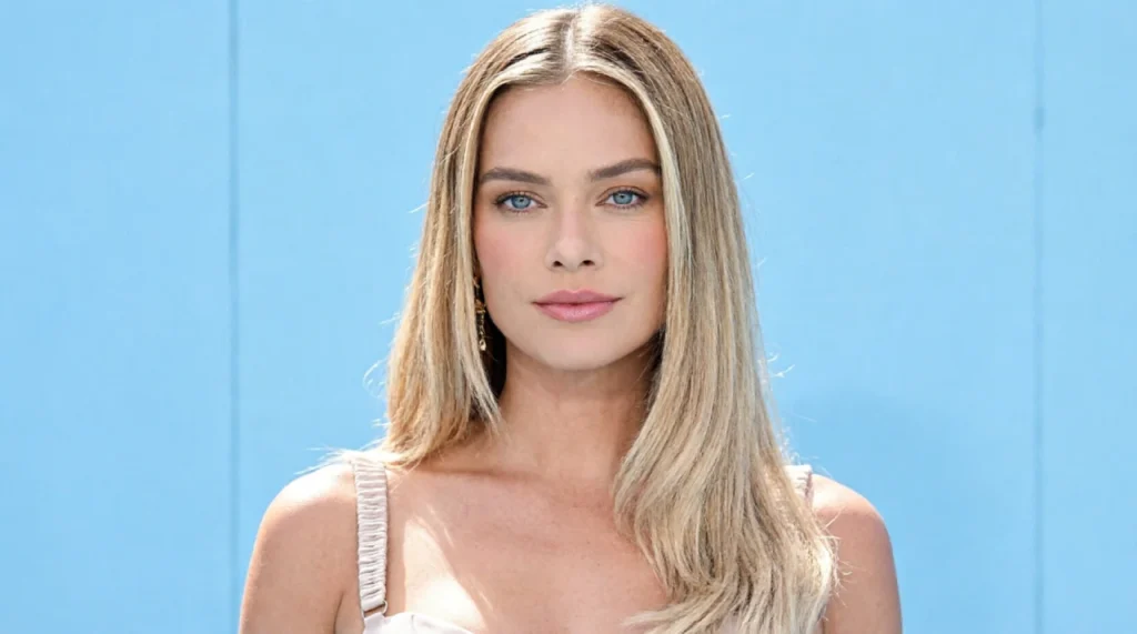Margot Robbie smiling warmly, blonde hair, in a white blazer.