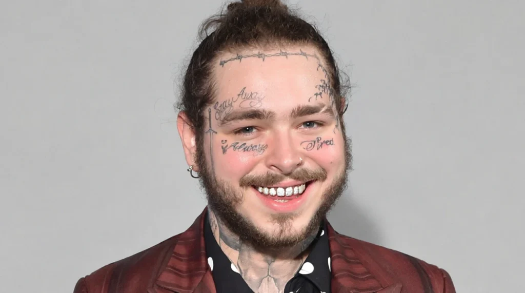 Post Malone with face tattoos, smiling slightly, in a hoodie.